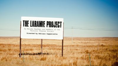 Image of white roadside billboard with black letters spelling out The Laramie Project set in landscape of yellow grass and blue skies